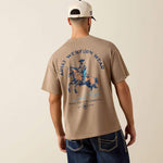 10079594 Ariat Mens Real Cowboy ToughStock Boxy T Shirt at boltranchstore.com free shipping in stock A man viewed from behind wearing a beige t-shirt with a blue and brown graphic of a cowboy on a horse, with 'ARIAT WESTERN WEAR' printed on the back. 