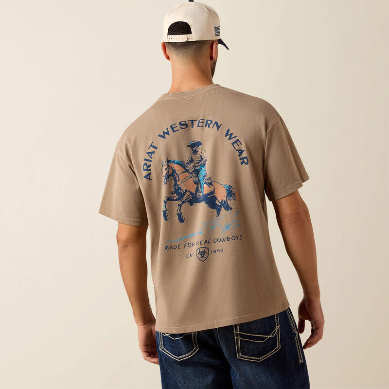 10079594 Ariat Mens Real Cowboy ToughStock Boxy T Shirt at boltranchstore.com free shipping in stock A man viewed from behind wearing a beige t-shirt with a blue and brown graphic of a cowboy on a horse, with 'ARIAT WESTERN WEAR' printed on the back. 