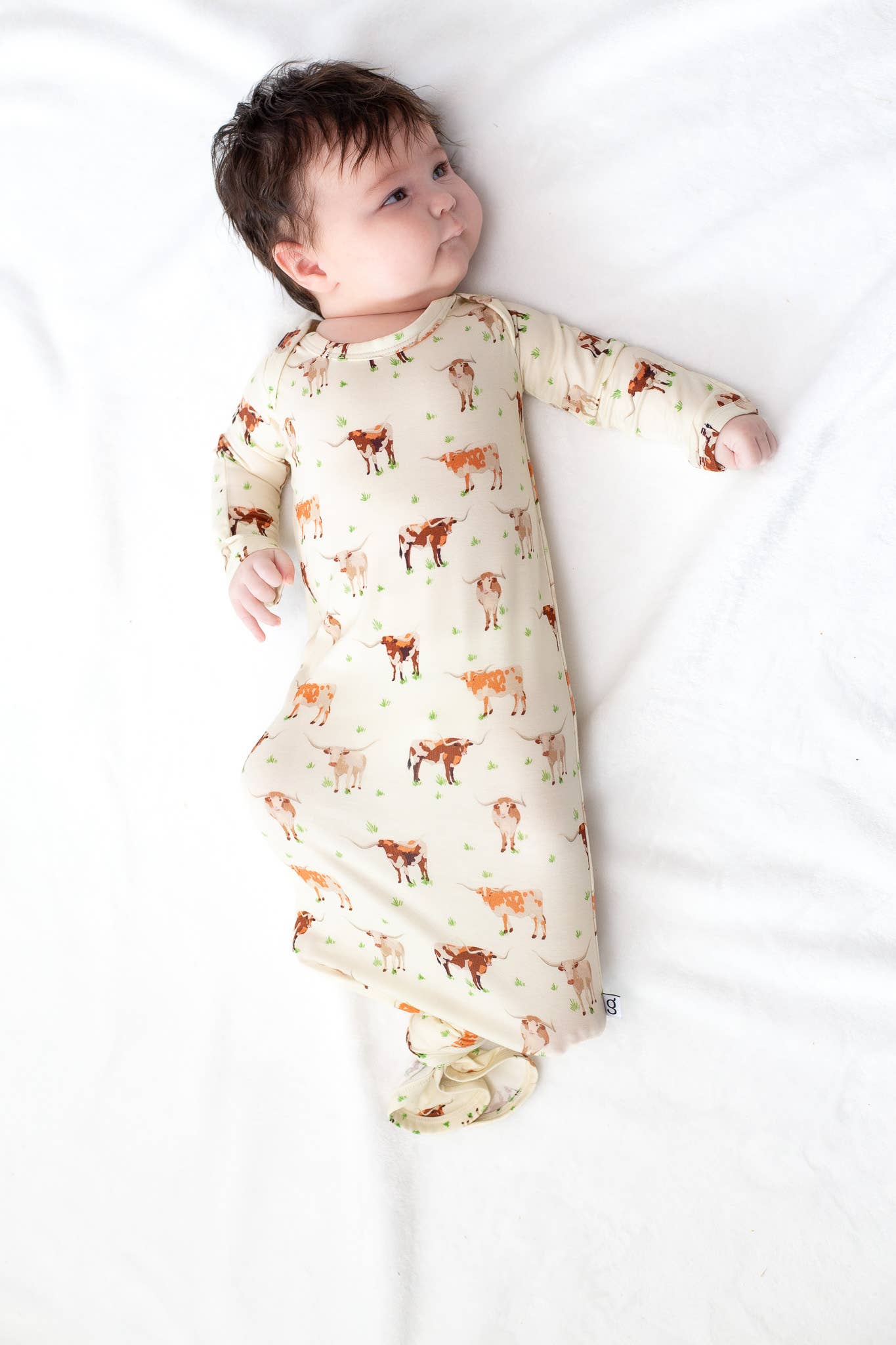 Baby lying on a white blanket wearing a long-sleeve onesie with animal prints. Longhorn Cows Knotted Infant Gown shop today at boltranchstore.com