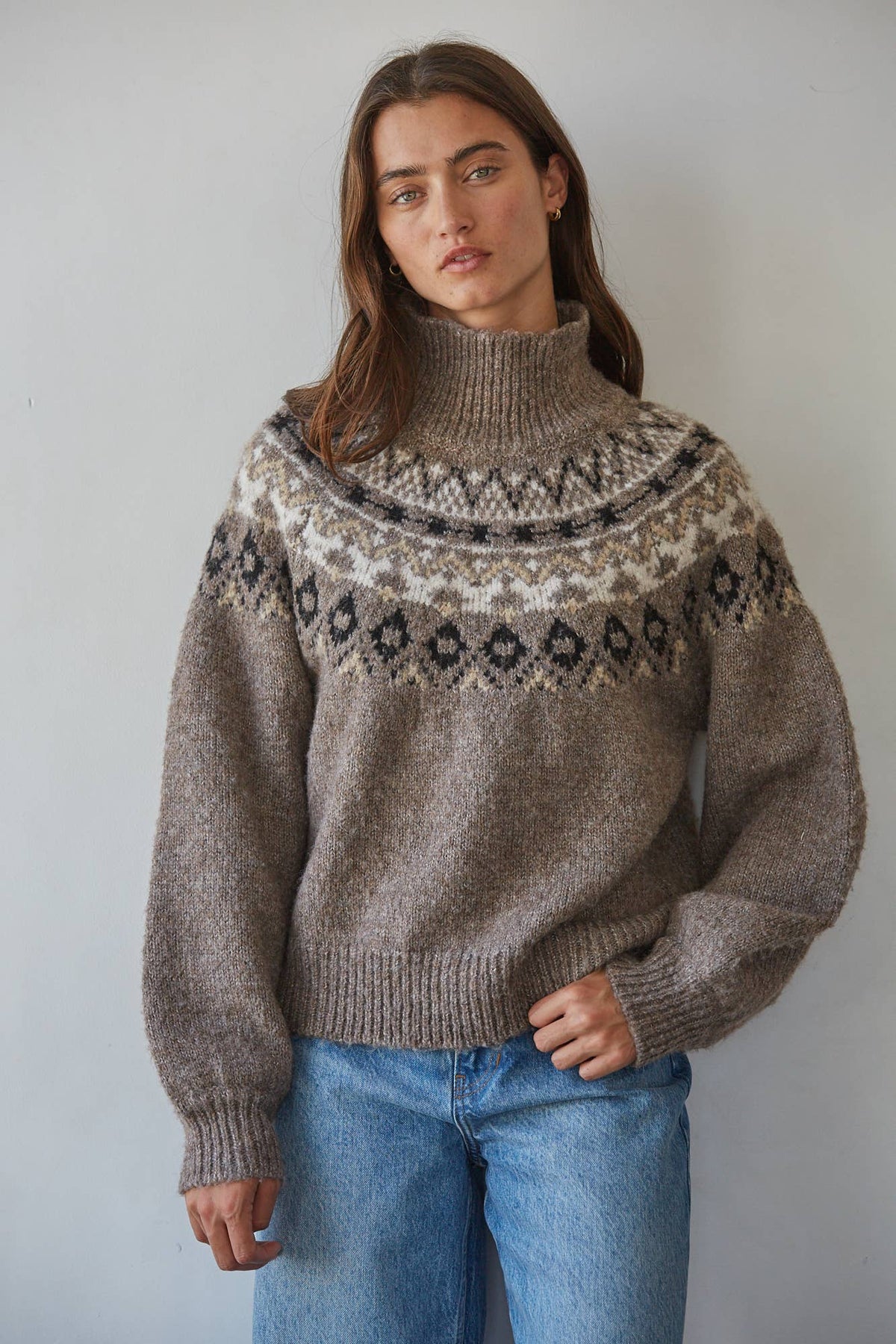 A woman wearing a brown Fair Isle knit turtleneck sweater with blue jeans, standing against a plain light gray wall.