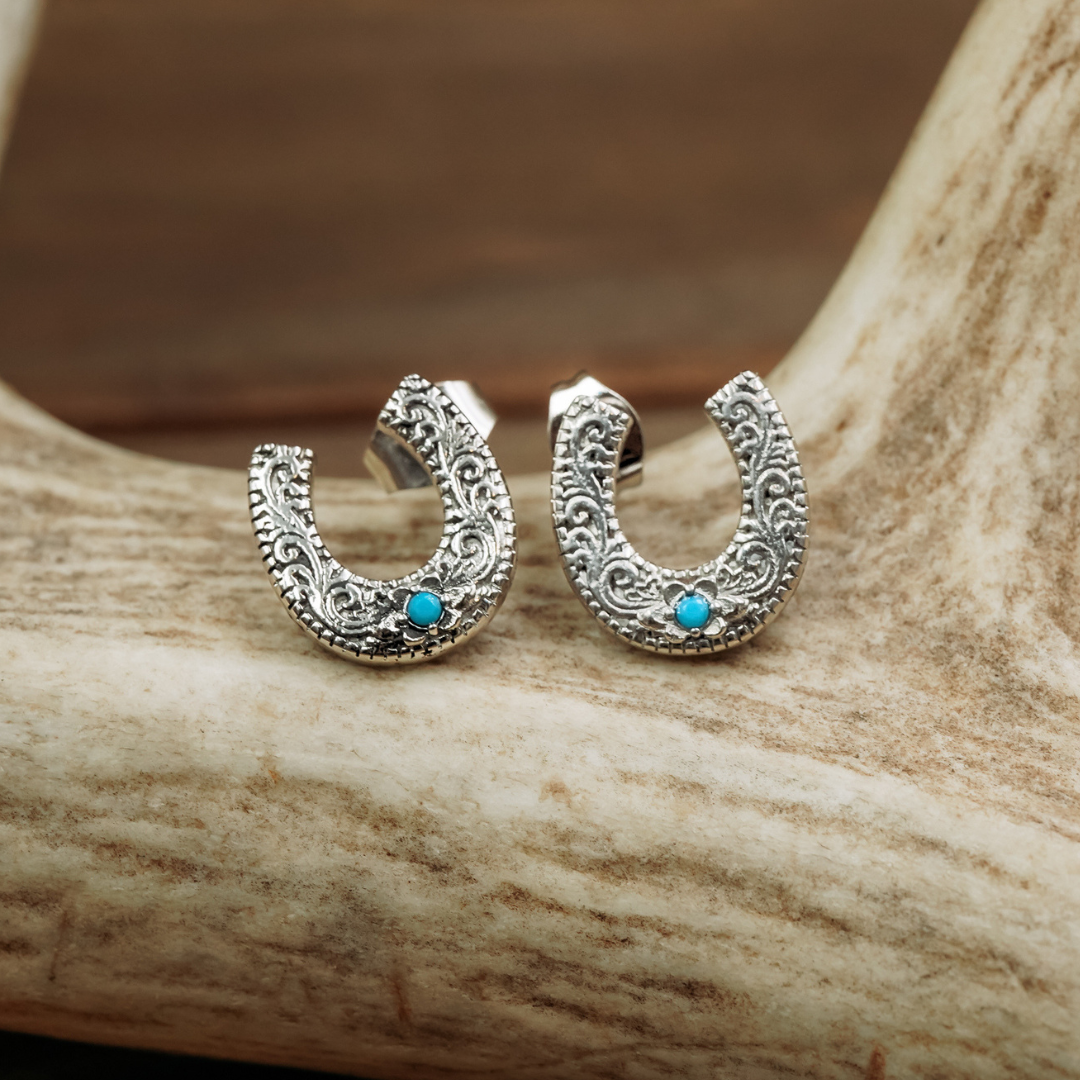Antler Ring The Lucky Studs at boltranchstore.com in stock free shipping A pair of silver crescent-shaped hoop earrings with intricate filigree detailing and a small turquoise stone, displayed on a light-colored antler against a warm brown background.