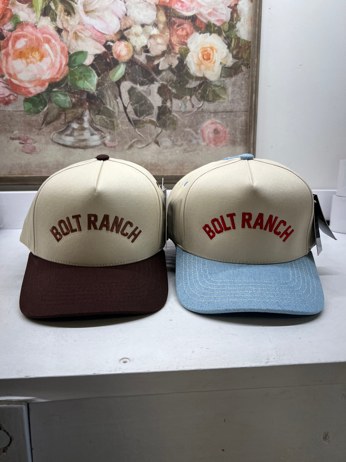 Two hats with 'Bolt Ranch' branding on a white surface, floral painting in the background. bolt ranch embroidered denim brim brown brim boltranchstore.com