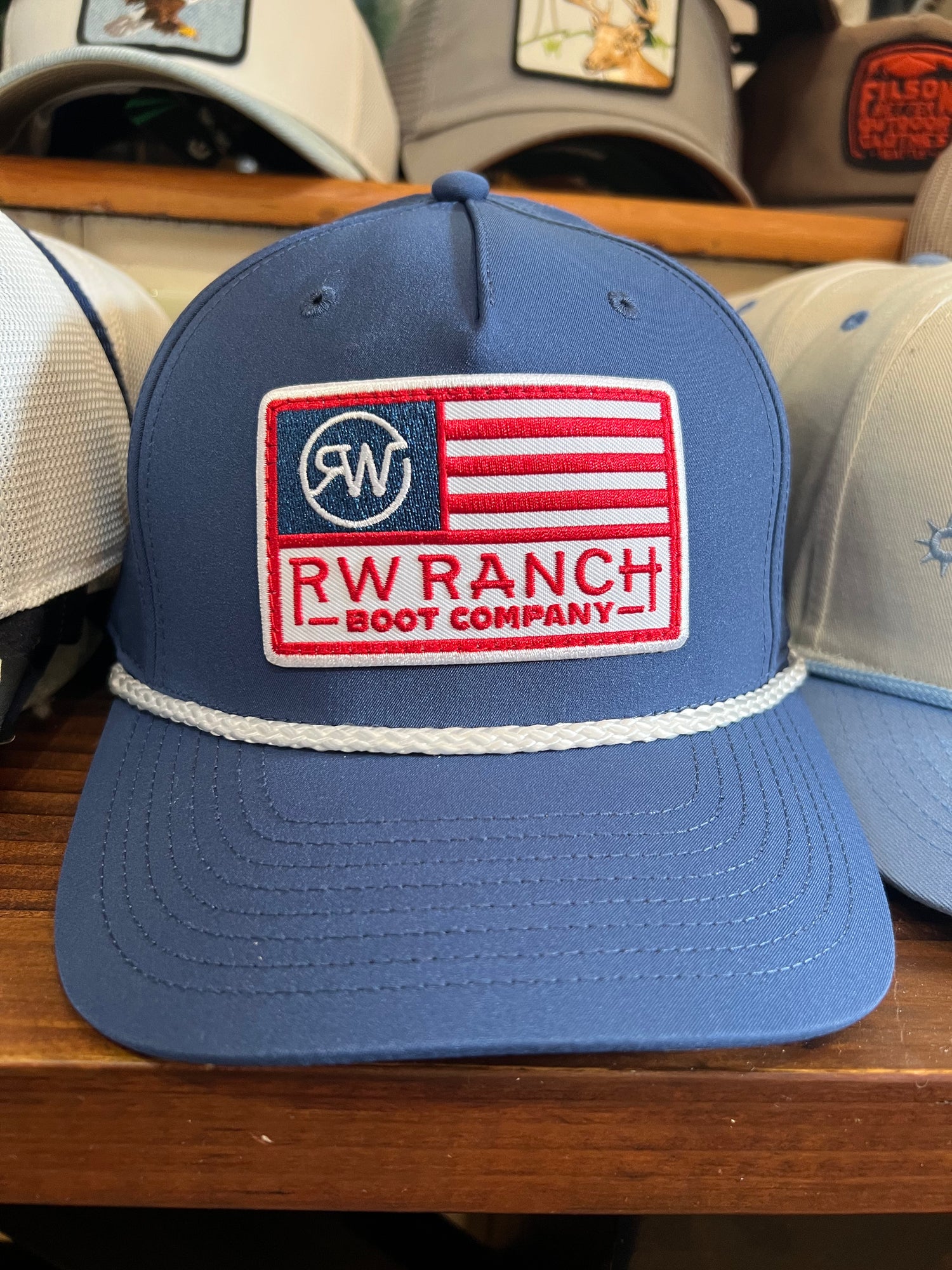 Blue cap with RW Ranch Boot Company logo on a wooden surface with american flag style logo red and white stripes r watson boltranchstore.com white rope detail