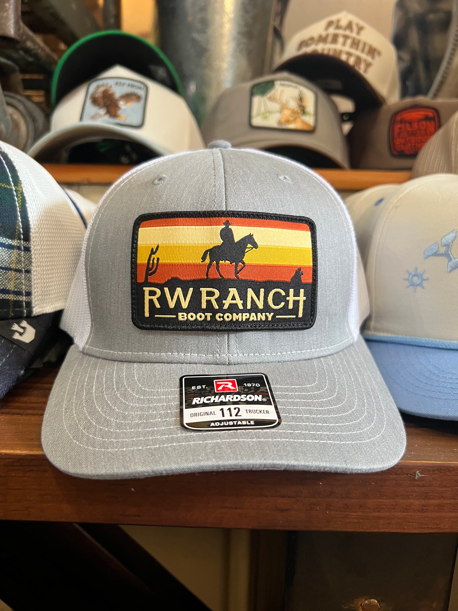 Gray cap with RW Ranch Boot Company logo with orange and yellow sunset and silhouette of cowboy on a horse. Trucker hat with white mesh on the back. R watson boltranchstore.com