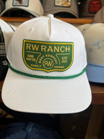 White cap with green and yellow 'RW Ranch' logo on a wooden surface with other caps in the background. R watson boltranchstore.com