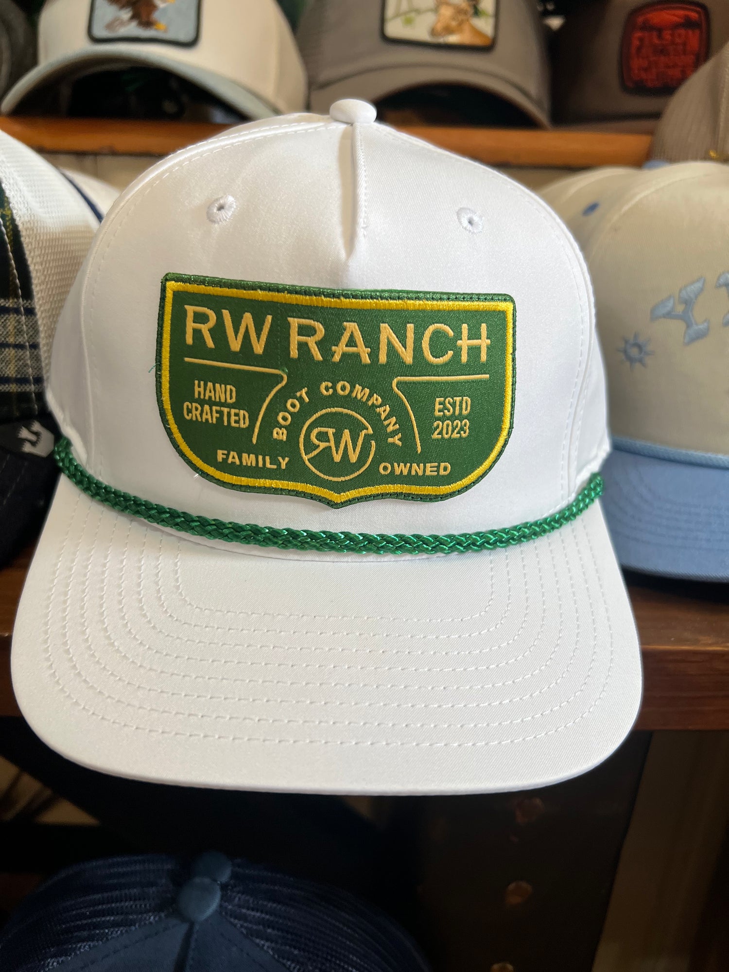 White cap with green and yellow 'RW Ranch' logo on a wooden surface with other caps in the background. R watson boltranchstore.com