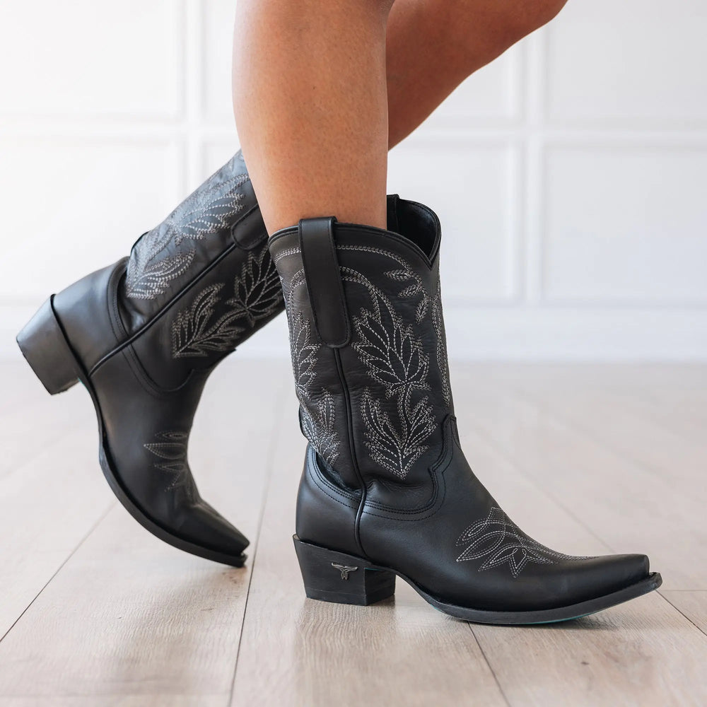 Lane Sandaga Midi Boot in Jet Black at boltranchhstore.com in stock free shipping Black cowboy boots with floral embroidery worn by a person on a light wooden floor.