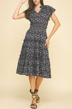 The Nadia navy floral dress features a charming ruffle collar and short sleeves, adorned with a delicate cream floral design. Its elastic waist ensures a comfortable fit, while the tiered skirt adds a graceful, flowing silhouette perfect for any occasion.