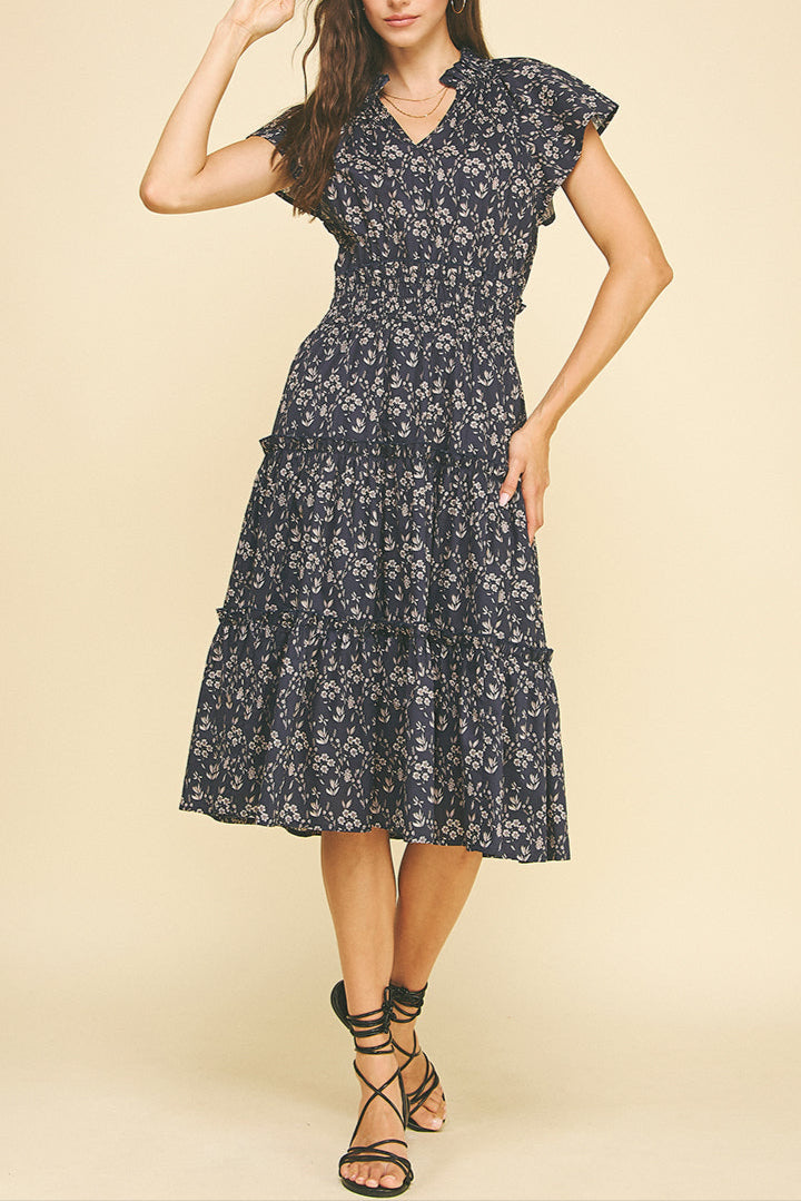 The Nadia navy floral dress features a charming ruffle collar and short sleeves, adorned with a delicate cream floral design. Its elastic waist ensures a comfortable fit, while the tiered skirt adds a graceful, flowing silhouette perfect for any occasion.
