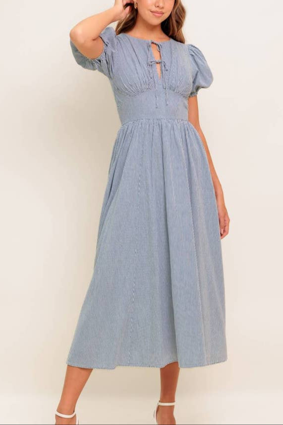 Nori Striped Dress This midi dress showcases a striped woven fabric with a round neckline and charming front bow ties. It features short puff sleeves, a fitted waist yoke, a flowing full skirt, and a convenient back zipper closure for easy wear.