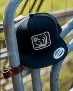 Red Dirt Hat features a deer antler patch on a black hat with black mesh back in a flat bill and snap back hat available at boltranchstore.com