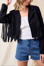Woman wearing a black fringe jacket, white top, and blue shorts with a beige hat against a neutral background