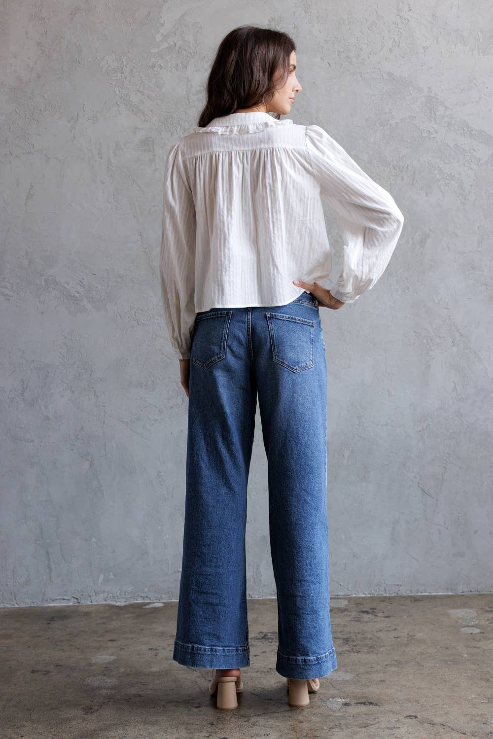 Person wearing a white blouse and blue jeans against a gray wall.